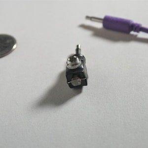 May include: A close-up of a small black audio adapter with a silver knob. The adapter has a 3.5mm jack on one end and a 1/4 inch jack on the other end.