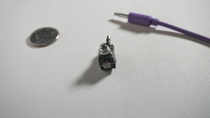 May include: A close-up of a small black audio adapter with a silver knob. The adapter has a 3.5mm jack on one end and a 1/4 inch jack on the other end.
