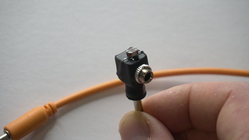 May include: A close-up of a black audio adapter with a silver 3.5mm jack. The adapter is connected to an orange cable.