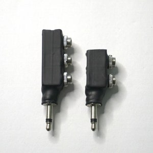 Może przedstawiać: Dwa czarne adaptery audio ze srebrnymi złączami i gniazdem 3,5 mm. Jeden adapter jest większy od drugiego. Adaptery znajdują się na białym tle.