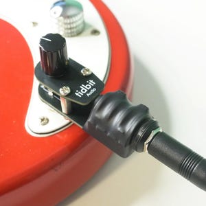 May include: A red electric guitar with a black audio adapter attached. The adapter has a black knob and the text "tidbit Audio" printed on it. A black audio cable is plugged into the adapter.