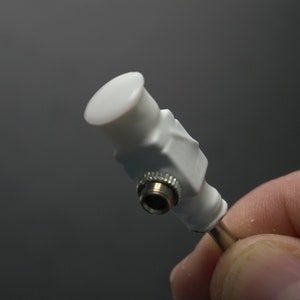 Pode incluir: Um conector de pl&aacute;stico branco com uma extremidade rosqueada prateada. O conector tem uma tampa redonda branca na parte superior.