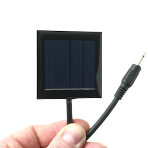 Könnte beinhalten: Ein kleines schwarzes Solarpanel mit einem schwarzen Kabel und einem Stecker. Das Panel ist etwa 5 cm breit und 5 cm hoch.
