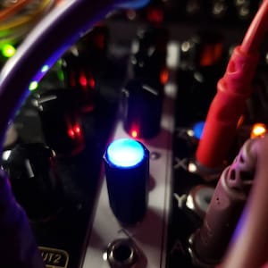 May include: Close-up of a black electronic music synthesiser with a blue LED light on a knob. The synthesiser has a variety of knobs and jacks.