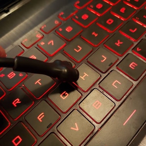 May include: A black laptop keyboard with red backlit keys. The keys are labeled with letters and numbers. A black tool is resting on the 'G' key.