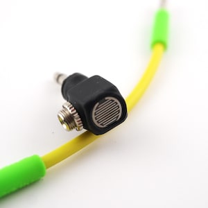 May include: A black, right-angled audio adapter with a silver threaded connector and a striped grill. It is connected to a yellow cable with green ends. The background is white.