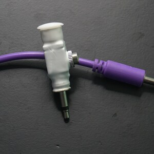 Pode incluir: Um cabo de &aacute;udio roxo com um conector branco e uma ponta prateada. O cabo est&aacute; conectado a um adaptador branco com um bot&atilde;o prateado.