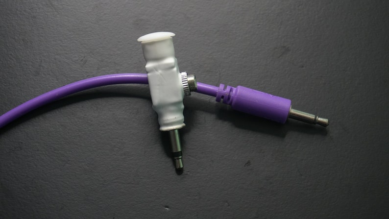 Pode incluir: Um cabo de &aacute;udio roxo com um conector branco e uma ponta prateada. O cabo est&aacute; conectado a um adaptador branco com um bot&atilde;o prateado.