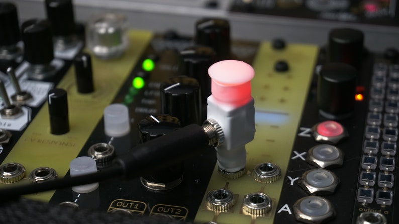 Pode incluir: Close-up de um sintetizador de m&uacute;sica eletr&oacute;nico branco e preto com um bot&atilde;o vermelho aceso. O sintetizador tem bot&otilde;es, interruptores e tomadas. O bot&atilde;o vermelho est&aacute; etiquetado como "A".