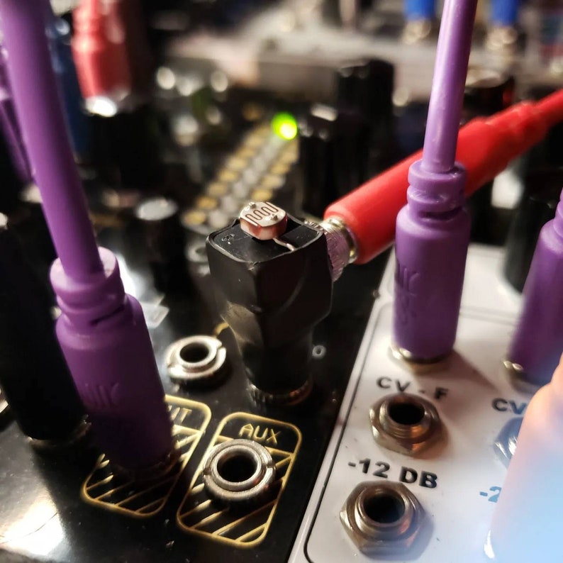 May include: Close-up of a black electronic module with purple and red cables plugged in. The module has a small silver resistor labeled "1000" and several jacks labeled "AUX", "CV-F", and "-12 DB".