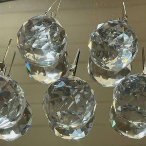 May include: Six clear crystal ball ornaments with faceted surfaces and silver-colored metal clips. The ornaments are arranged in a close-up shot, reflecting light and creating a sparkling effect. These decorative ornaments are suitable for holiday or home decor.