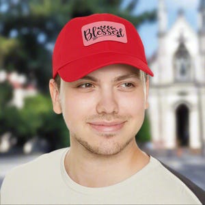 Könnte beinhalten: Eine leuchtend rote Baseballkappe mit gebogenem Schirm. Auf der Kappe befindet sich ein rechteckiger Aufnäher mit dem Wort "Blessed" in Schreibschrift, begleitet von einem floralen Design. Die Kappe wird von einer Person getragen.