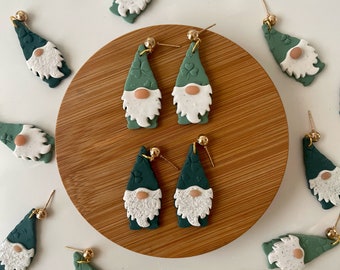 May include: Handmade green gnome earrings with white beards and pink noses. The earrings have gold-colored posts and are arranged on a wooden surface. Some earrings feature a shamrock design on the hat. The earrings are a festive accessory.