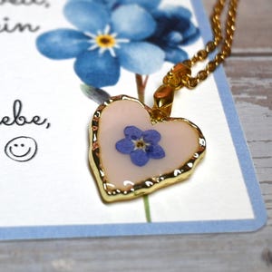 May include: A heart-shaped pendant necklace with a gold chain. The pendant features a dried blue flower encased in resin, framed by a gold border. The necklace rests on a card with German text and a blue floral design.