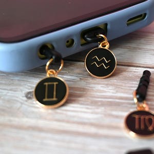 Puede incluir: Una funda de teléfono azul claro con colgantes dorados con símbolos del zodiaco. Los colgantes son negros con contornos dorados y están unidos a la toma de audio del teléfono. También se ven otros colgantes con símbolos del zodiaco.