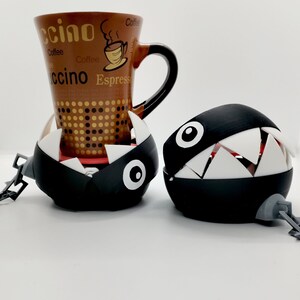 May include: Two black and white 3D printed  'Boo'  character  coasters from the Super Mario Bros. video game. The coasters have a chain attached to them. One coaster is holding a brown coffee mug with the word 'ccino' on it.