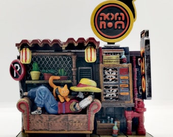 Puede incluir: Un diorama en miniatura de una pequeña tienda con un techo rojo, un letrero amarillo que dice "Kofi Nom" y una figura sentada en un sofá con un gato. La tienda tiene una ventana con un ventilador y una boca de incendios roja.