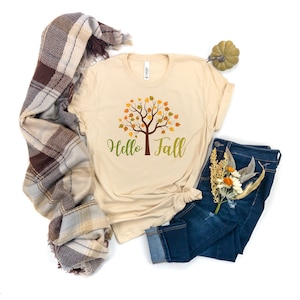 Peut inclure: Un t-shirt beige avec un arbre vert aux feuilles orange et jaunes et le texte "Hello Fall" imprimé sur le devant.