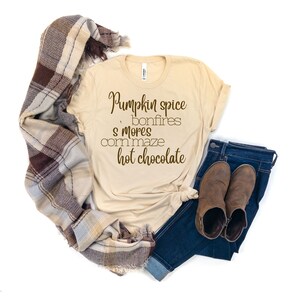 May include: A beige t-shirt with the text "Pumpkin spice, bonfires, s'mores, corn maze, hot chocolate" printed in brown on the front. The shirt is styled with a brown and beige plaid scarf, blue jeans, and brown ankle boots.