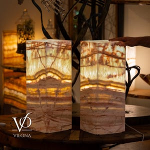 May include: Two rectangular lamps made of translucent banded onyx stone with warm internal illumination. The stone features natural horizontal layers of cream, beige, and reddish-brown colors. A white power cord extends from the base of the lamps.