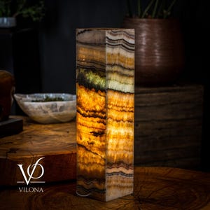 May include: A rectangular onyx lamp with a warm, glowing interior. The lamp features layers of tan, brown, and white tones, creating a natural, striped pattern. The base is a solid, rectangular shape. The lamp is lit, showcasing the stone's translucence.