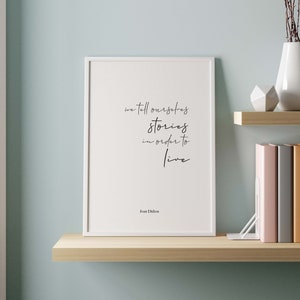 Puede incluir: Una impresión enmarcada en blanco con una cita que dice "we tell ourselves stories in order to live" de Joan Didion. La impresión está en un estante con un jarrón de ramas secas, una escultura geométrica y una pila de libros.