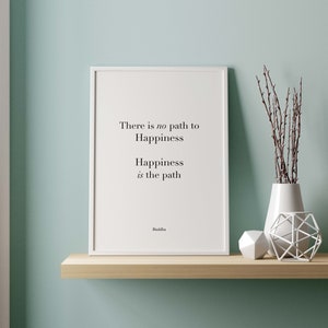 Op de afbeelding: Een witte ingelijste print met een citaat van Boeddha: "Er is geen pad naar geluk. Geluk is het pad." De print staat op een houten plank met een witte vaas en een wit geometrisch object.