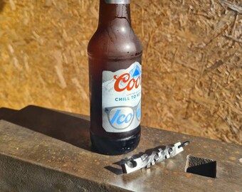 Può includere: Una bottiglia di birra in vetro marrone con un'etichetta blu e bianca che dice "Cool Chill to Relax" e un apribottiglie argentato su una superficie metallica.