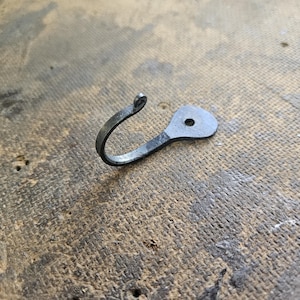 Könnte beinhalten: Handgeschmiedeter Metallhaken mit gebogenem Design und kleinem, kreisförmigem Loch. Der Haken ist dunkelgrau und scheint aus Eisen oder Stahl zu bestehen. Er wird wahrscheinlich zum Aufhängen von Gegenständen verwendet. Auf einer strukturierten, braunen Oberfläche.