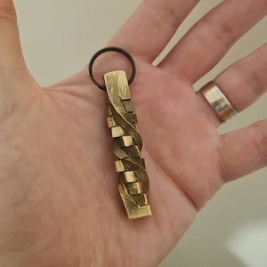 Puede incluir: Un llavero de metal de color dorado con un diseño trenzado. El llavero tiene un simple anillo en la parte superior para sujetar las llaves.
