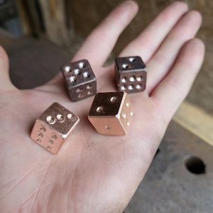 May include: Four metal dice, two copper and two bronze, resting in a hand. Each die features the standard arrangement of circular indentations, representing the numbers one to six. The dice are small, approximately 1.5 cm per side.