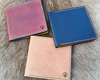 Op de afbeelding: Drie leren portemonnees in verschillende kleuren: bruin, blauw en beige. Elke portemonnee heeft een gestikte rand en een klein logo in het midden.