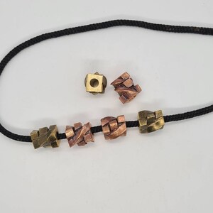 Op de afbeelding: Een zwarte koordketting met verschillende geometrische kralen in brons- en kopertinten. De kralen hebben een textuur, kubusachtig ontwerp. Twee extra kralen worden apart getoond, één kubusvormig en één met een meer abstracte vorm.