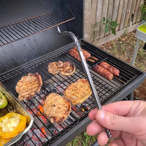 Könnte beinhalten: Ein schwarzer Grill mit verschiedenen Speisen, die darauf gegrillt werden, darunter Schweinekoteletts, Würstchen und Gemüse. Der Grill ist im Freien aufgebaut und im Vordergrund ist eine Hand zu sehen, die eine Zange hält.