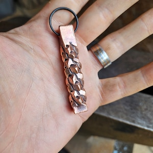 Puede incluir: Un llavero de cobre con un diseño trenzado. El llavero tiene un anillo de metal negro en la parte superior.