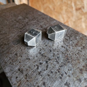 Puede incluir: Dos dados de metal plateado con los números 1 y 2 en ellos. Los dados están sobre una superficie de metal.