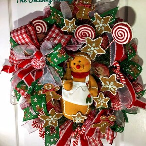 May include: A Christmas wreath decorated with gingerbread men, candy canes, red and white plaid ribbon, and a gingerbread woman holding a rolling pin. The wreath is made on a green base and has a burlap bow.