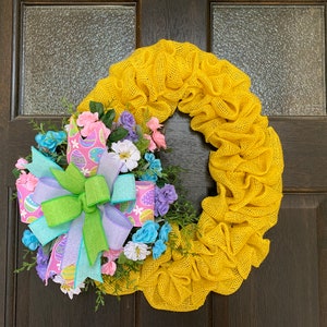 May include: A yellow burlap wreath with a colorful bow and floral accents. The bow features ribbons in pastel colors and Easter egg patterns. The wreath is decorated with pink, blue, and white flowers, creating a festive spring decoration.