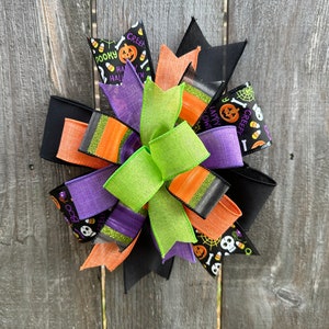 May include: A Halloween-themed bow with black, orange, green, and purple ribbons. The ribbons have various Halloween-themed prints, including pumpkins, candy corn, and skulls. The bow is tied with a black ribbon.