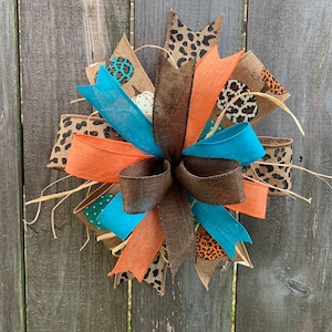 May include: A decorative bow featuring a mix of burlap, leopard print, and solid ribbons in teal, orange, and brown. Accented with faux straw and pumpkin-themed fabric, it's a festive fall decoration.