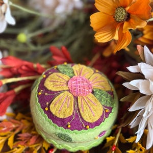 May include: A round, green pincushion with a felt flower design. The flower has yellow petals, a pink center, and a purple base. The pincushion is surrounded by colorful fall foliage, including orange, red, and white flowers.