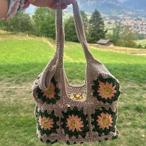 Può includere: Una borsa all'uncinetto color beige con un motivo a quadrati. Ogni quadrato presenta un fiore giallo al centro, circondato da petali rosa chiaro e un bordo verde scuro. La borsa è dotata di due manici integrati e un'apertura superiore.