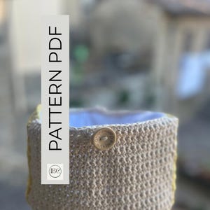 Può includere: Un cestino all'uncinetto di forma quadrata color crema chiaro con un bordo giallo. Sulla parte anteriore è fissato un piccolo bottone rotondo in legno chiaro con l'incisione "Handmade with love". Un'etichetta verticale riporta la scritta "PATTERN PDF".