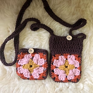 Può includere: Due borse all'uncinetto in filato marrone scuro con un motivo floreale quadrato in rosa, giallo e arancione ruggine. Ogni borsa ha una chiusura con bottone in legno chiaro. Una lunga tracolla all'uncinetto marrone scuro unisce le borse.