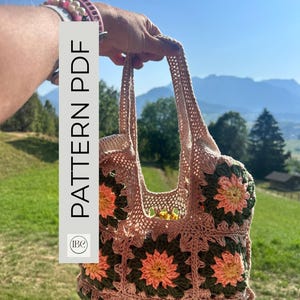 Può includere: Una borsa all'uncinetto realizzata con piastrelle granny unite, caratterizzata da un motivo floreale nei toni del rosa polvere, verde oliva scuro e giallo. La borsa ha due manici e una base in filato rosa chiaro. Il testo PATTERN PDF è visibile a sinistra.