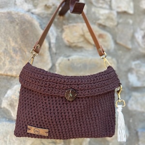 Può includere: Una borsa a mano all'uncinetto marrone scuro con patta ripiegabile fissata da un singolo bottone rotondo scuro. La borsa include un cinturino in pelle color cuoio fissato con chiusure in metallo dorato e una piccola nappa bianca laterale.