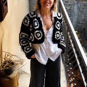 Può includere: Un cardigan all'uncinetto bianco e nero con un motivo ripetitivo a piastrelle granny, indossato sopra una camicia chiara abbottonata e pantaloni in denim scuro a gamba larga. Il cardigan ha maniche lunghe e una lunghezza corta con lavorazione a maglia.