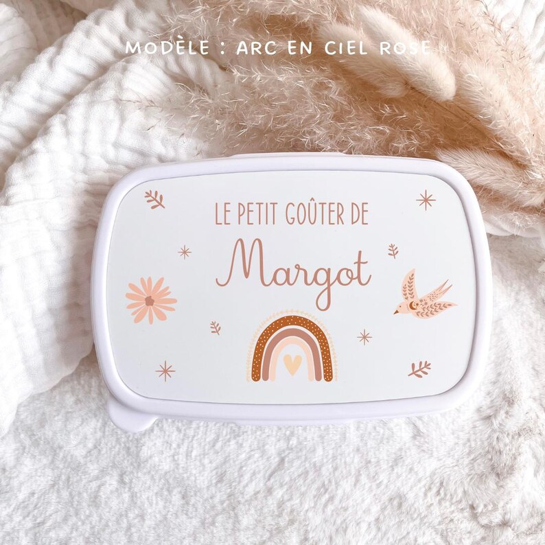 May include: A white rectangular lunch box featuring the text LE PETIT GO&Ucirc;TER DE Margot in brown lettering. The lid is decorated with a brown and pink rainbow, a small bird, a flower, and star accents. The design is printed on a smooth white surface.