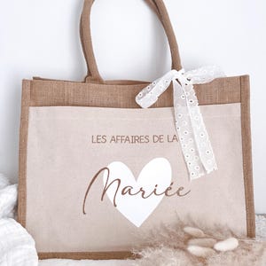 May include: A brown burlap tote bag with a white heart and the text "Les Affaires de la Mari&eacute;e" in brown lettering. The bag has a white lace ribbon tied around the handles.