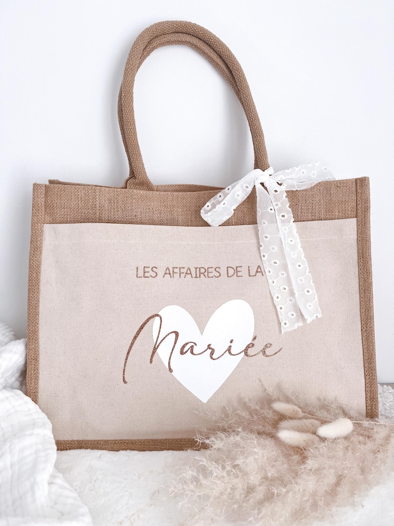 May include: A brown burlap tote bag with a white heart and the text "Les Affaires de la Mari&eacute;e" in brown lettering. The bag has a white lace ribbon tied around the handles.